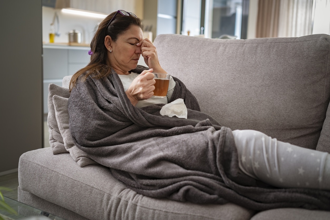 Woman resting on sofa drinking hot tea and massaging her nose